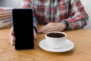 Elinde kahve fincanı ve kitaplarla ahşap masada oturan akıllı telefon ekranı gösteren tanınmamış bir adam..