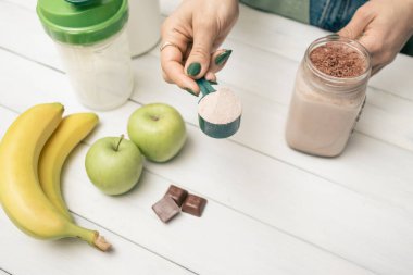 Kot pantolonlu ve gömlekli genç bir kadın, elinde protein tozlu kaşık, cam kavanoz protein içeceği kokteyli, beyaz ahşap masa üstünde milkshake veya smoothie, çikolata parçaları, muz, elma.