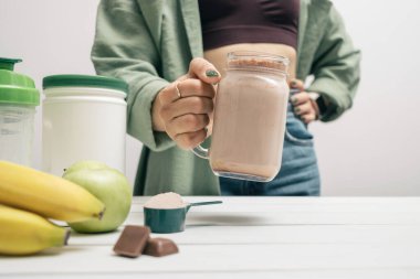 Kot pantolonlu ve gömlekli genç bir kadın elinde cam kavanoz protein içeceği kokteyli, milkshake veya smoothie ile beyaz ahşap masa üzerinde ölçüm kaşığı protein tozu, çikolata parçaları, muz ve elma..