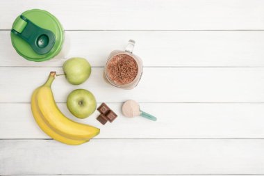 Cam kavanoz protein içeceği kokteyli, milkshake veya smoothie, protein tozuyla ölçme kaşığı, çikolata parçaları, muz ve elmalar beyaz ahşap masa üzerinde, üst görünüm.