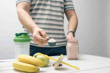 Elinde protein tozlu kaşık tutan adam, cam kavanoz protein içeceği kokteyli, milkshake veya smoothie beyaz ahşap masa üzerinde çikolata parçaları, muz, elma.