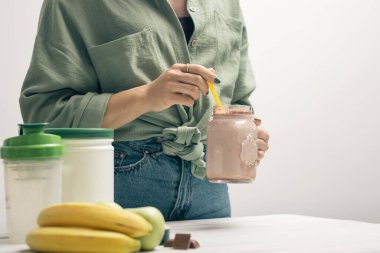 Kot pantolonlu ve gömlekli genç bir kadın elinde cam kavanoz protein içeceği kokteyli, milkshake veya smoothie ile beyaz ahşap masa üzerinde ölçüm kaşığı protein tozu, çikolata parçaları, muz ve elma..