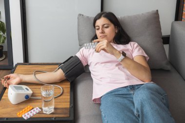 Evdeki kanepede uzanmış genç bir kadın modern dijital tonometreyle elinde haplarla kan basıncını ölçüyor..