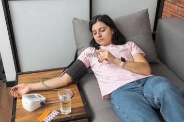 Evdeki kanepede uzanmış genç bir kadın modern dijital tonometreyle elinde haplarla kan basıncını ölçüyor..