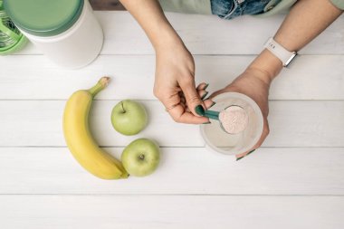 Elinde ölçü kaşığı olan genç bir kadın beyaz ahşap masada muz ve elmalarla süt tozu tozu karıştırıyor. Protein içeceği yapma süreci, üst görünüm.