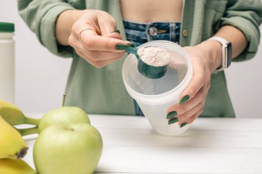 Elinde kaşık olan kot pantolonlu ve gömlekli genç bir kadın muz ve elmalarla beyaz ahşap masada süt tozunun bir kısmını karıştırıyor. Protein içeceği yapma süreci.