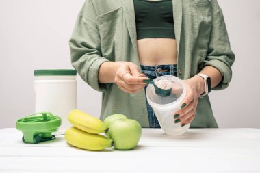 Elinde kaşık olan kot pantolonlu ve gömlekli genç bir kadın muz ve elmalarla beyaz ahşap masada süt tozunun bir kısmını karıştırıyor. Protein içeceği yapma süreci.