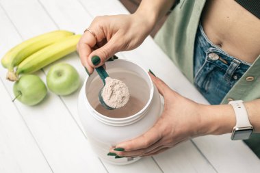 Kot pantolonlu ve tişörtlü bir kadın elinde portakal suyu içeren kaşık, plastik kavanozun üstünde beyaz ahşap masa üzerinde muz ve elma meyveli protein tozu tutuyor. Protein içeceği yapma süreci.