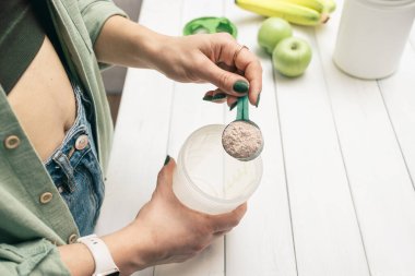 Elinde kaşık olan kot pantolonlu ve gömlekli genç bir kadın muz ve elmalarla beyaz ahşap masada süt tozunun bir kısmını karıştırıyor. Protein içeceği yapma süreci.