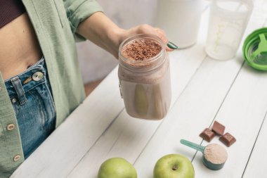 Kot pantolonlu ve gömlekli genç bir kadın elinde cam kavanoz protein içeceği kokteyli, milkshake veya smoothie ile beyaz ahşap masa üzerinde ölçüm kaşığı protein tozu, çikolata parçaları, muz ve elma..