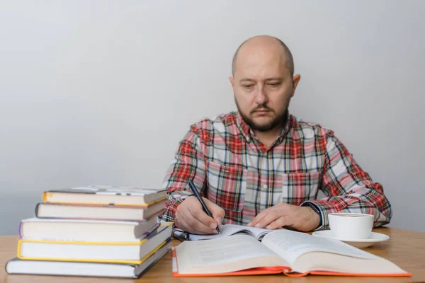 Man writing in notepad, reading book, studying or working, sitting at the table with stack of books and cup of coffee, focus on the hand with pencil.