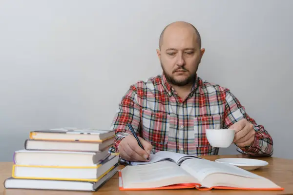 Man writing in notepad, reading book, studying or working, sitting at the table with stack of books and holding cup of coffee. focus on the hand with pencil.