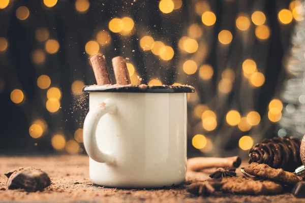 Homemade spicy hot chocolate drink with cinnamon stick, star anise, grated chocolate in enamel mug on dark background with cookies, cacao powder and chocolate pieces, Christmas lights bokeh.
