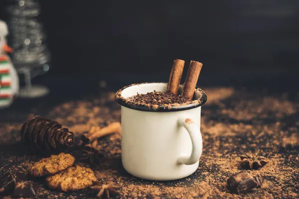 Homemade spicy hot chocolate drink with cinnamon stick, star anise, grated chocolate in enamel mug on dark background with cookies, cacao powder and chocolate pieces.