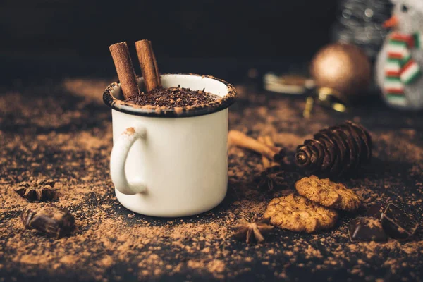 Homemade spicy hot chocolate drink with cinnamon stick, star anise, grated chocolate in enamel mug on dark background with cookies, cacao powder and chocolate pieces.