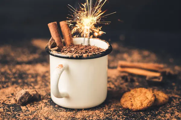 Homemade spicy hot chocolate drink with cinnamon stick, star anise, grated chocolate and sparklers in enamel mug on dark background with cookies, cacao powder and chocolate pieces.