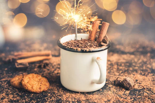 Homemade hot chocolate drink with cinnamon stick, star anise, grated chocolate and sparklers in enamel mug on dark background with cookies, cacao powder and chocolate pieces, Christmas lights bokeh.