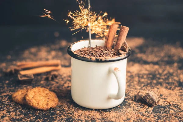 Homemade spicy hot chocolate drink with cinnamon stick, star anise, grated chocolate and sparklers in enamel mug on dark background with cookies, cacao powder and chocolate pieces.