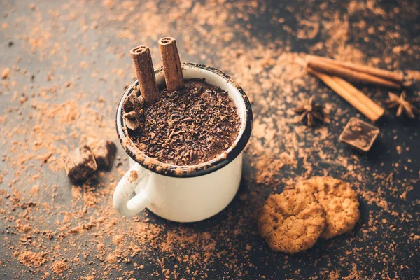 Homemade spicy hot chocolate drink with cinnamon stick, star anise, grated chocolate in enamel mug on dark background with cookies, cacao powder and chocolate pieces.