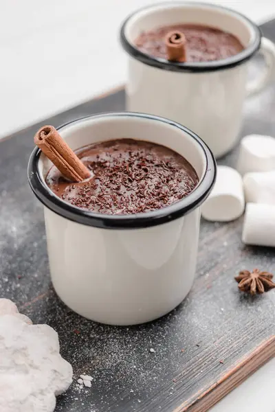 Homemade spicy hot chocolate drink with cinnamon stick, grated chocolate in enamel cup on wooden table with cookies, white marshmallows and star anise.