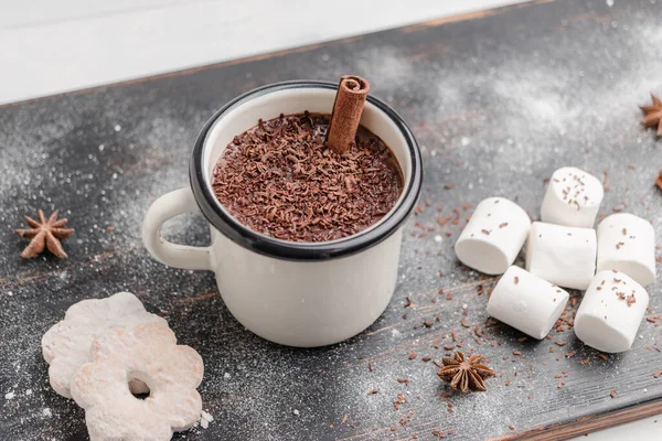 Homemade spicy hot chocolate drink with cinnamon stick, grated chocolate in enamel cup on wooden table with cookies, white marshmallows and star anise.