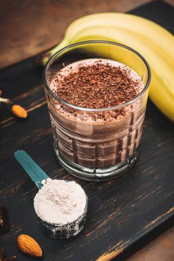 Milkshake smoothie, koyu ahşap tahta üzerinde rendelenmiş çikolatalı protein içeceği muz, ölçüm kaşığında protein tozu, badem, tarçın çubuğu, çikolata parçaları..