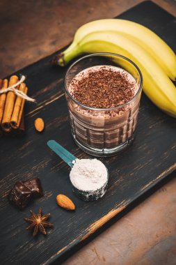 Milkshake smoothie, koyu ahşap tahta üzerinde rendelenmiş çikolatalı protein içeceği muz, ölçme kaşığında protein tozu, yıldız anason, badem, tarçın çubuğu, çikolata parçaları..