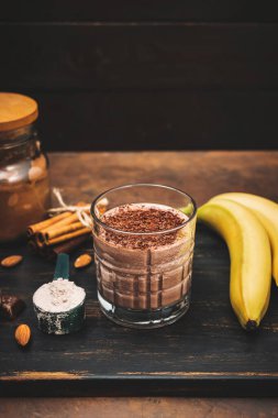 Milkshake smoothie, koyu ahşap tahta üzerinde rendelenmiş çikolatalı protein içeceği muz, ölçme kaşığında protein tozu, yıldız anason, badem, tarçın çubuğu, çikolata parçaları..