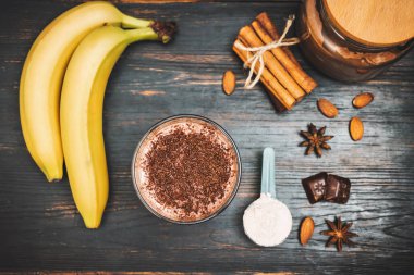 Milkshake smoothie, koyu ahşap bir bardakta rendelenmiş çikolatalı protein içeceği muz, ölçme kaşığında protein tozu, yıldız anason, badem, tarçın çubuğu, çikolata parçaları, üst manzara..