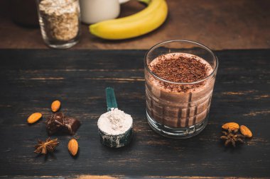 Milkshake smoothie, koyu ahşap tahta üzerinde rendelenmiş çikolatalı protein içeceği muz, ölçme kaşığında protein tozu, yıldız anason, badem, tarçın çubuğu, çikolata parçaları..