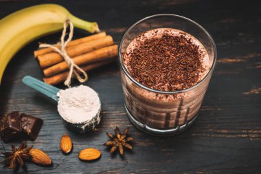 Milkshake smoothie, koyu ahşap tahta üzerinde rendelenmiş çikolatalı protein içeceği muz, ölçme kaşığında protein tozu, yıldız anason, badem, tarçın çubuğu, çikolata parçaları..
