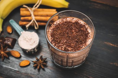 Milkshake smoothie, koyu ahşap tahta üzerinde rendelenmiş çikolatalı protein içeceği muz, ölçme kaşığında protein tozu, yıldız anason, badem, tarçın çubuğu, çikolata parçaları..