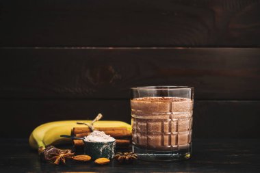 Milkshake smoothie, koyu ahşap tahta üzerinde rendelenmiş çikolatalı protein içeceği muz, ölçme kaşığında protein tozu, yıldız anason, badem, tarçın çubuğu, çikolata parçaları..