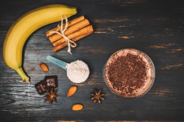 Milkshake smoothie, koyu ahşap bir bardakta rendelenmiş çikolatalı protein içeceği muz, ölçme kaşığında protein tozu, yıldız anason, badem, tarçın çubuğu, çikolata parçaları, üst manzara..