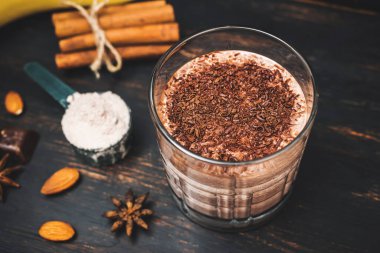 Milkshake smoothie, koyu ahşap tahta üzerinde rendelenmiş çikolatalı protein içeceği muz, ölçme kaşığında protein tozu, yıldız anason, badem, tarçın çubuğu, çikolata parçaları..