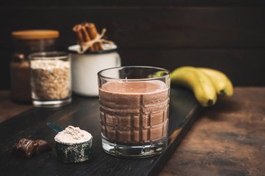 Çikolatalı milkshake smoothie, koyu ahşap tahta üzerinde bir bardakta protein içeceği muz, ölçü kaşığında protein tozu ve çikolata parçaları..