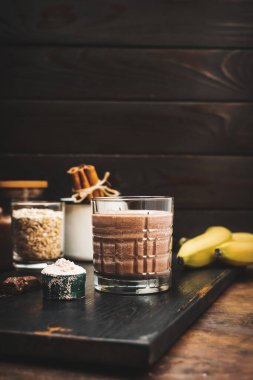 Çikolatalı milkshake smoothie, koyu ahşap tahta üzerinde bir bardakta protein içeceği muz, ölçü kaşığında protein tozu ve çikolata parçaları..