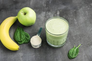 Bardakta yeşil detoks smoothie, protein tozuyla ölçme kaşığı, ıspanak yapraklarından karışık vejetaryen içeceği, koyu arkaplanda elma ve muz meyvesi, sağlıklı beslenme konsepti..