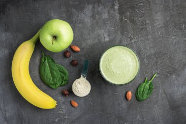 Bardakta yeşil detoks smoothie, protein tozuyla ölçme kaşığı, ıspanak yapraklarından karıştırılmış vejetaryen içeceği, koyu arkaplanda elma ve muz meyvesi, sağlıklı beslenme konsepti, üst görüş..