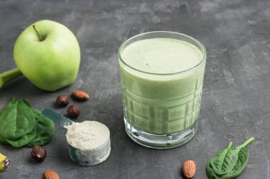 Bardaktaki yeşil detoks smoothie, protein tozuyla ölçme kaşığı, ıspanak yaprakları ve arkaplanda elma meyvesinden karıştırılmış vejetaryen içeceği, sağlıklı beslenme konsepti..