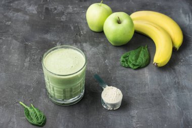 Bardakta yeşil detoks smoothie, protein tozuyla ölçme kaşığı, ıspanak yapraklarından karışık vejetaryen içeceği, koyu arkaplanda elma ve muz meyvesi, sağlıklı beslenme konsepti..