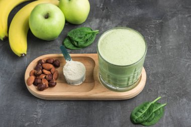 Bardakta yeşil detoks smoothie, protein tozuyla ölçme kaşığı, ıspanak yapraklarından karışık vejetaryen içeceği, koyu arkaplanda elma ve muz meyvesi, sağlıklı beslenme konsepti..