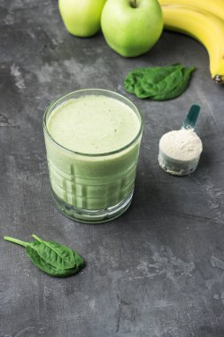 Bardakta yeşil detoks smoothie, protein tozuyla ölçme kaşığı, ıspanak yapraklarından karışık vejetaryen içeceği, koyu arkaplanda elma ve muz meyvesi, sağlıklı beslenme konsepti..