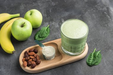 Bardakta yeşil detoks smoothie, protein tozuyla ölçme kaşığı, ıspanak yapraklarından karışık vejetaryen içeceği, koyu arkaplanda elma ve muz meyvesi, sağlıklı beslenme konsepti..