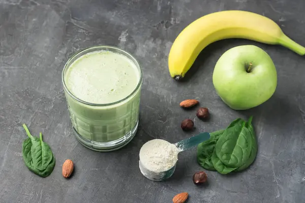 Bardakta yeşil detoks smoothie, protein tozuyla ölçme kaşığı, ıspanak yapraklarından karışık vejetaryen içeceği, koyu arkaplanda elma ve muz meyvesi, sağlıklı beslenme konsepti..