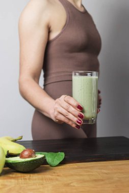 Spor kıyafetli, elinde sağlıklı yeşil vejetaryen meyve püresi bardağıyla tanınmayan atletik bir kadın. muz, ıspanak, elma ve avokado bir masada..