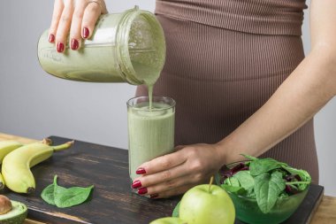 Spor kıyafetleriyle tanınmayan atletik kadın bardağa sağlıklı yeşil vejetaryen içeceği döküyor. muz, ıspanak, elma ve avokado bir masada..