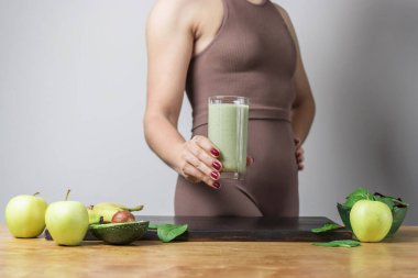 Spor kıyafetli, elinde sağlıklı yeşil vejetaryen meyve püresi bardağıyla tanınmayan atletik bir kadın. muz, ıspanak, elma ve avokado bir masada..