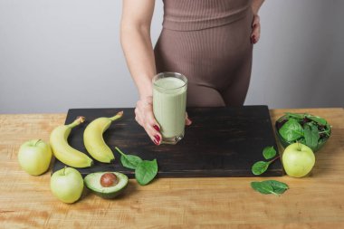 Spor kıyafetli, elinde sağlıklı yeşil vejetaryen meyve püresi bardağıyla tanınmayan atletik bir kadın. muz, ıspanak, elma ve avokado bir masada..