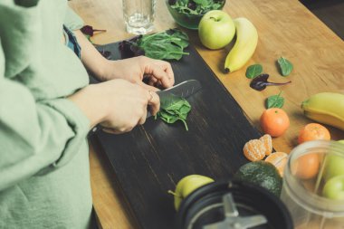 Yeşil ıspanak yaprakları kesen bir kadın, sağlıklı vejetaryen smoothie hazırlarken ıspanak, muz, elma ve avokado karıştırıcıyı masaya koyar..